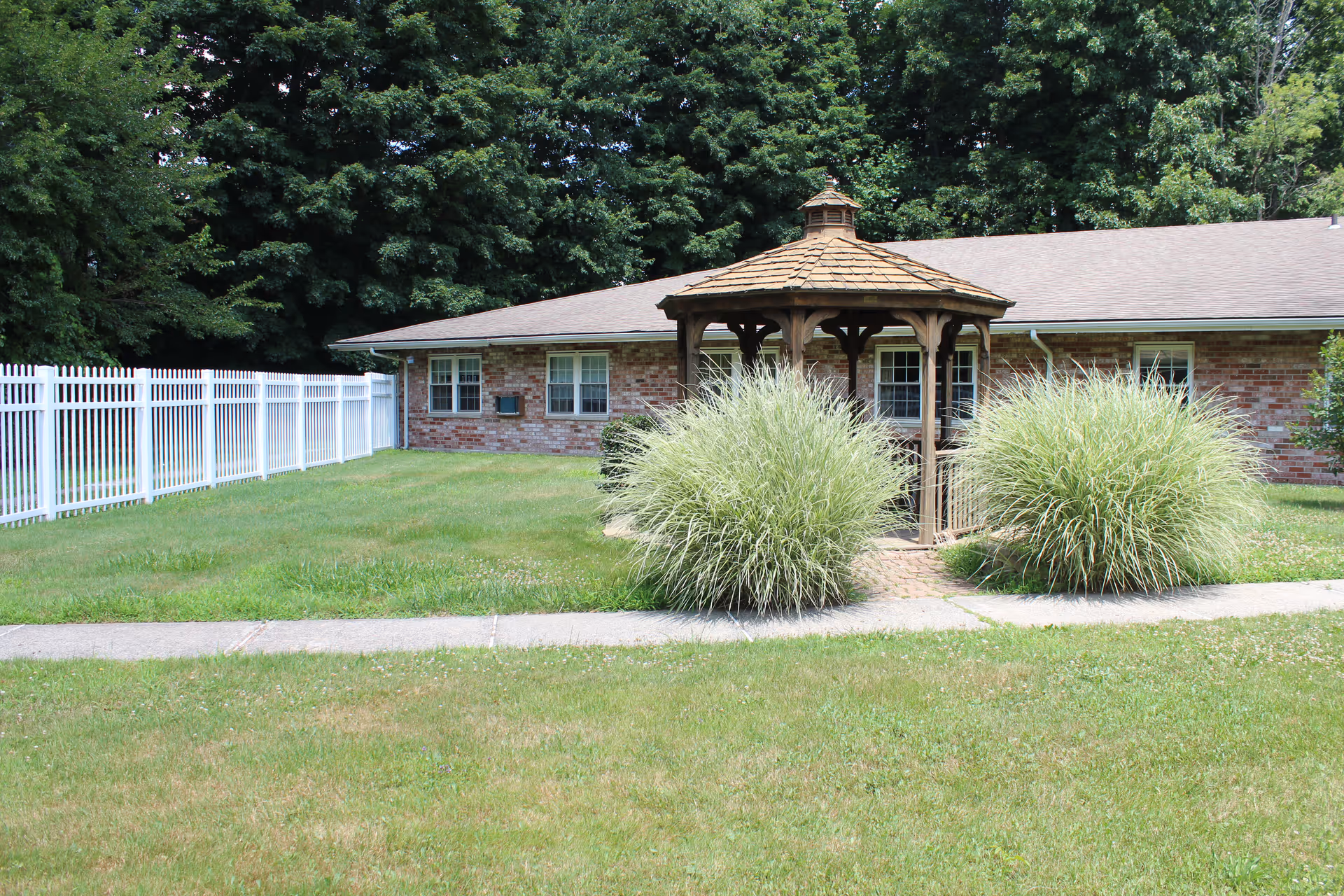 A grassy outdoor area with a wooden gazebo surrounded by two large ornamental grass bushes. Behind the gazebo is a single-story brick building with several windows. A white fence runs along the left side of the grassy area, and tall green trees form the background.