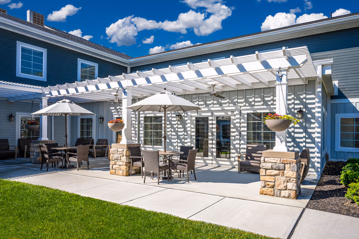 Outdoor patio area with a white pergola, tables and umbrellas, wicker chairs, and hanging flower planters in front of a gray building.
