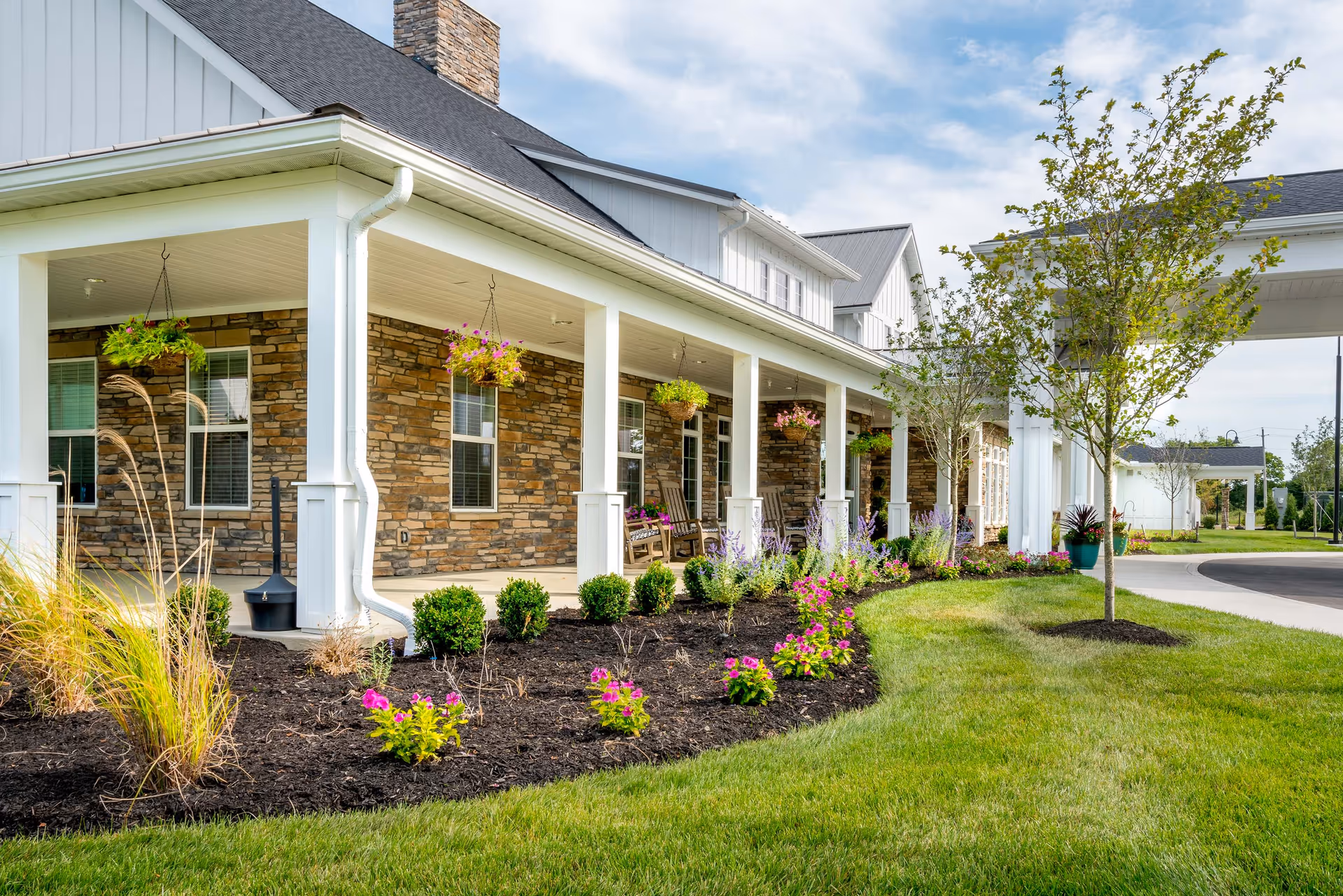 Exterior view of a senior living facility with a covered porch featuring hanging flower baskets, stone and white siding walls, landscaped garden beds with colorful flowers and shrubs, and a curved driveway with green grass and small trees.