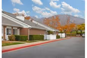 Exterior view of a single-story brick building with white trim, surrounded by a paved driveway and landscaped with bushes and trees with autumn foliage. Mountains are visible in the background under a partly cloudy sky.