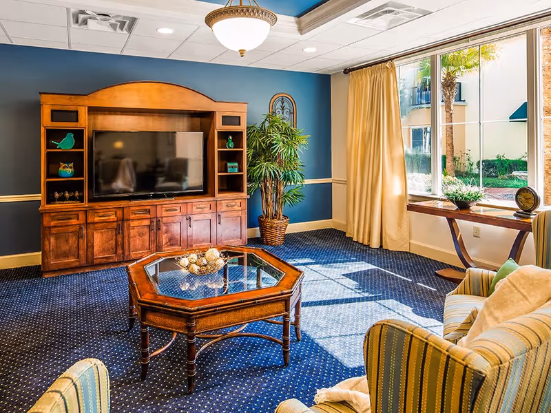 A cozy living room with a large wooden entertainment center holding a flat-screen TV, a glass-top wooden coffee table, striped upholstered armchairs, a potted plant, and a large window with beige curtains letting in natural light.
