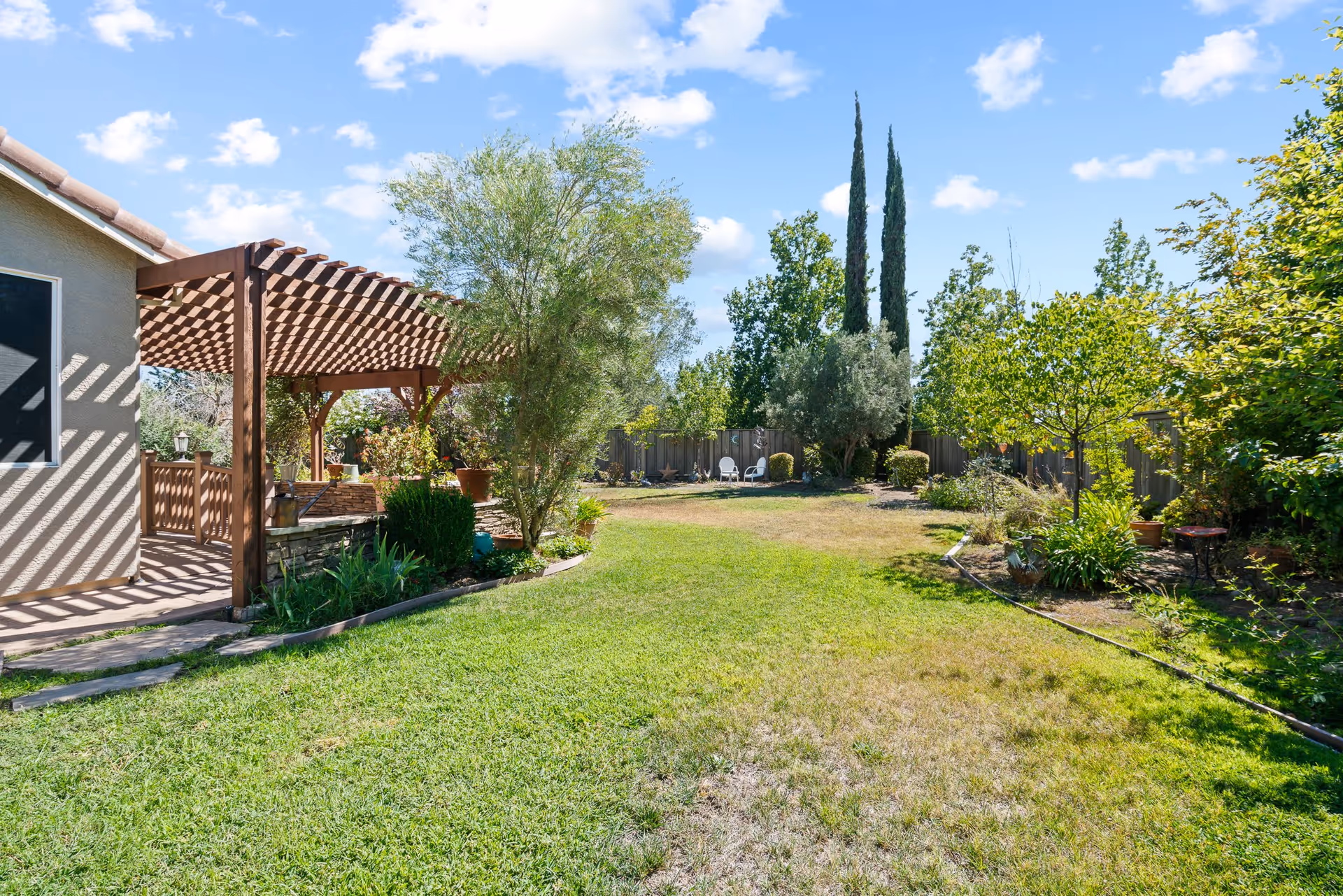 A sunny backyard garden area with a green lawn, various trees and shrubs, a wooden pergola attached to the side of a house, and two white chairs near a wooden fence in the background under a blue sky with scattered clouds.