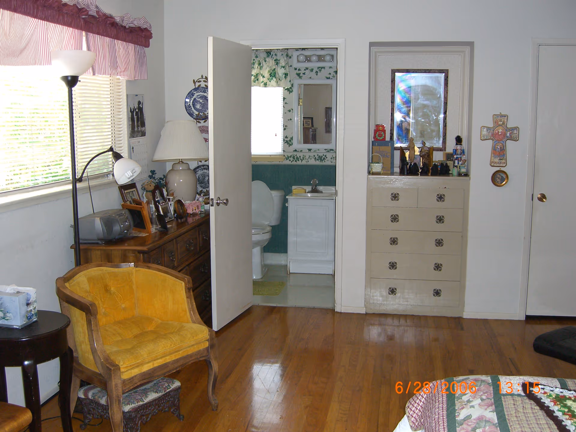 Interior view of a residential room with wooden flooring, a yellow upholstered chair, a wooden dresser with a lamp and photo frames, and an open door leading to a bathroom with a toilet and sink. The room has a window with blinds and a red and white valance, a floor lamp, and a chest of drawers with decorative items and a framed picture above it.