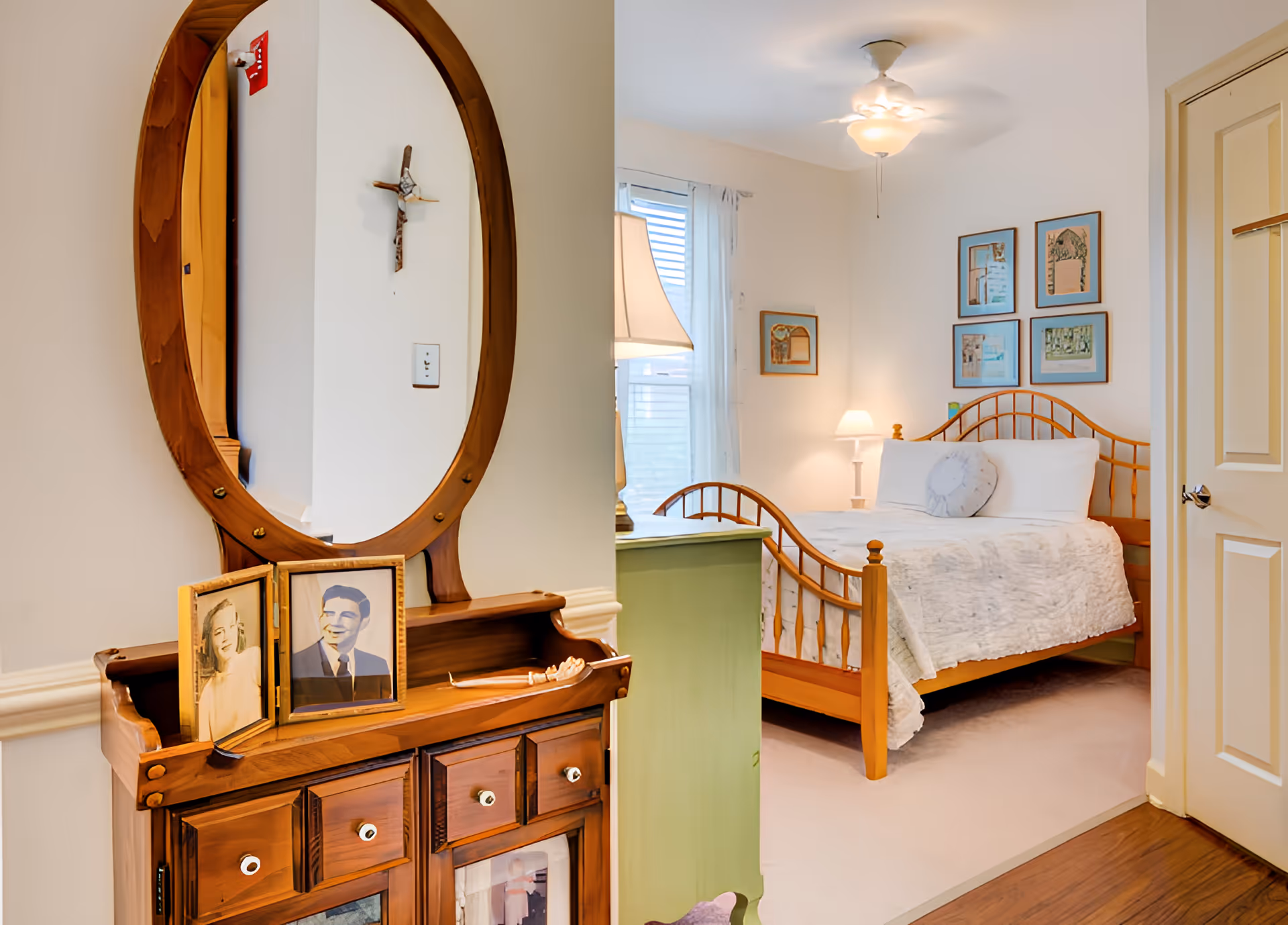 A cozy bedroom with a wooden bed frame, white bedding, and a round decorative pillow. The room features a ceiling fan with a light, framed pictures on the wall, a window with sheer curtains, and a bedside lamp on a green nightstand. In the foreground, there is a wooden dresser with an oval mirror and framed black-and-white photographs.
