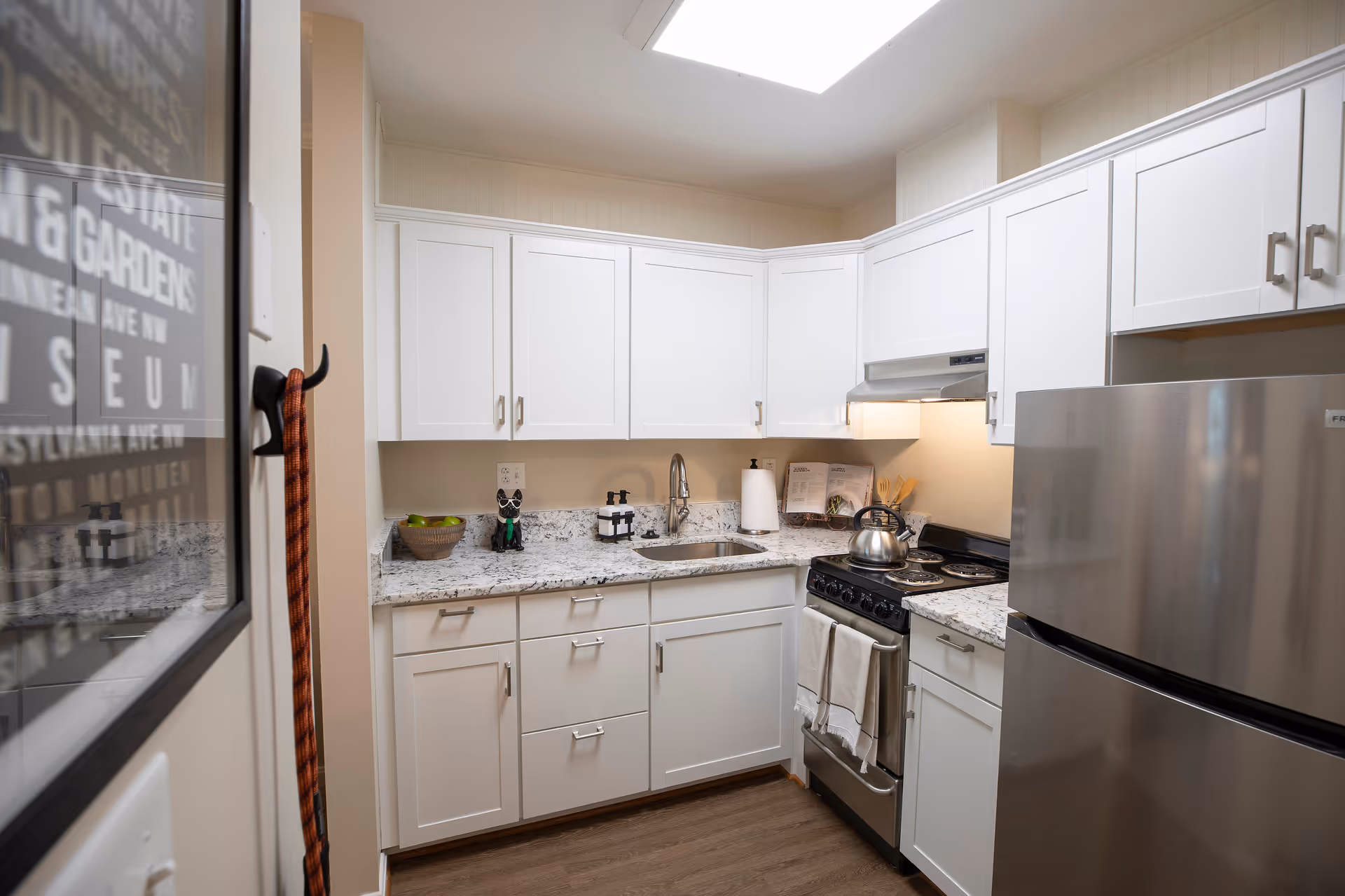 A compact kitchen with white cabinets, granite countertops, a stainless steel refrigerator, a stove with a kettle on it, a sink, and various kitchen items including a bowl of green apples and soap dispensers. There is a skylight above providing natural light.
