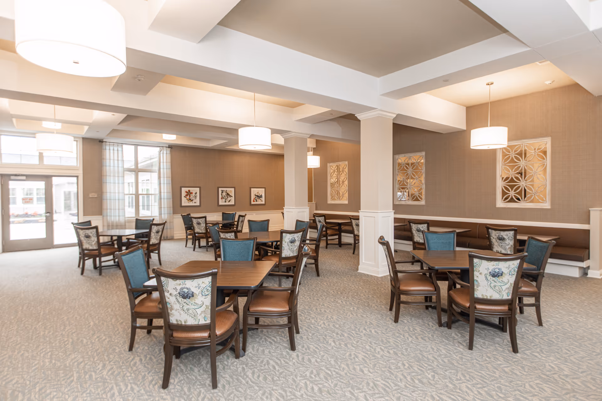 A spacious dining room in a senior living facility with multiple wooden tables and chairs arranged neatly. The chairs have floral patterned backs and brown seats. The room features beige walls with decorative framed artwork and mirrors, large windows with sheer curtains, and modern pendant lights hanging from the ceiling. The carpet has a subtle leaf pattern.