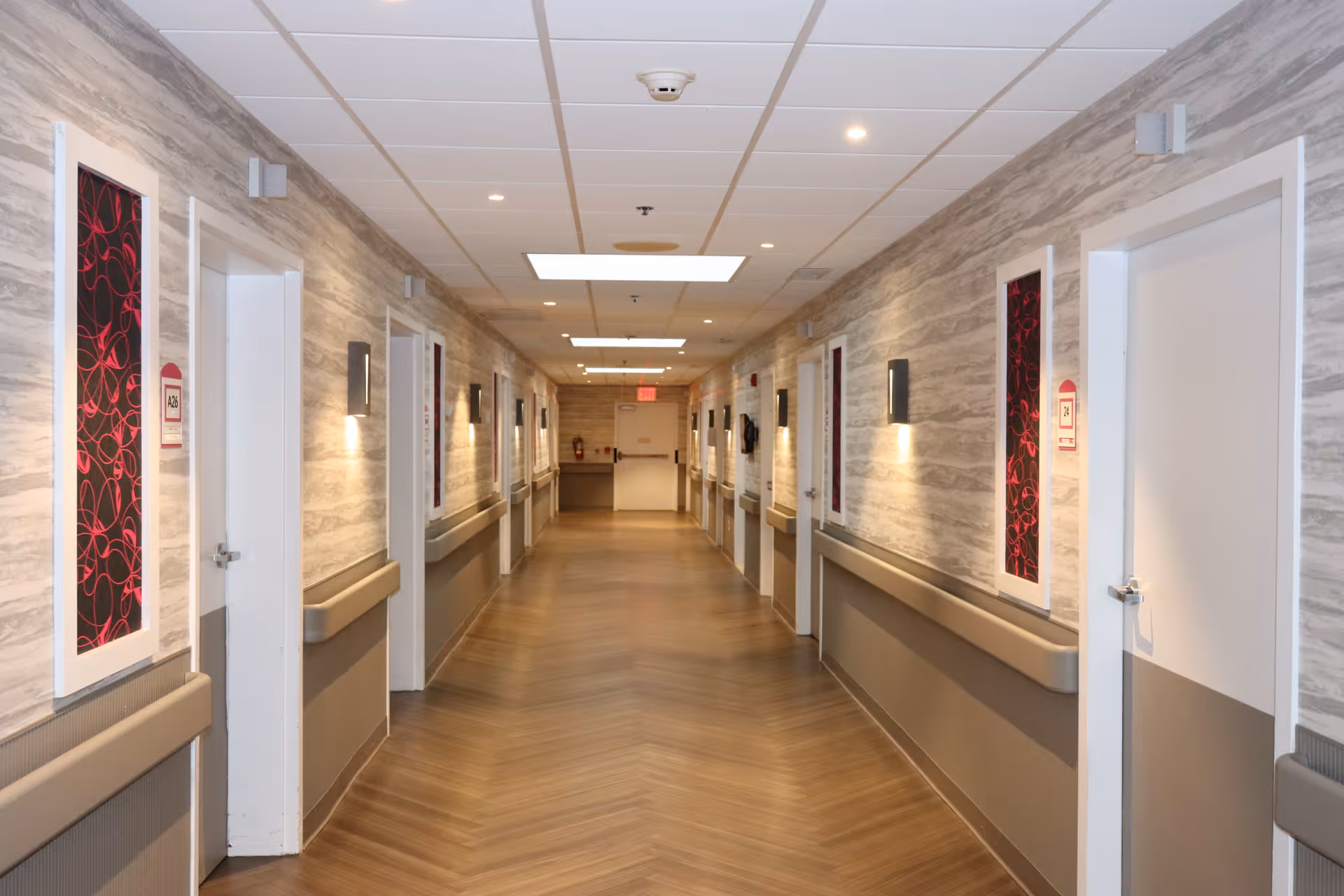 A well-lit, long hallway in a senior living facility with multiple closed white doors on both sides. The walls have a textured, light-colored design with handrails running along both sides. There are modern wall-mounted lights and decorative panels with red and black patterns between the doors. The ceiling has recessed lighting and a smoke detector, and the floor has a wood-patterned finish.