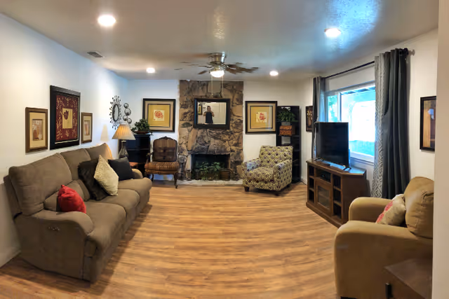 A cozy living room with wooden flooring, a stone fireplace in the center, and a ceiling fan above. The room has a brown sofa with multiple cushions on the left, a patterned armchair and a wooden chair near the fireplace, and a beige armchair on the right. A flat-screen TV is placed on a wooden TV stand next to a large window with dark curtains. The walls are decorated with framed artwork and a decorative wall clock.