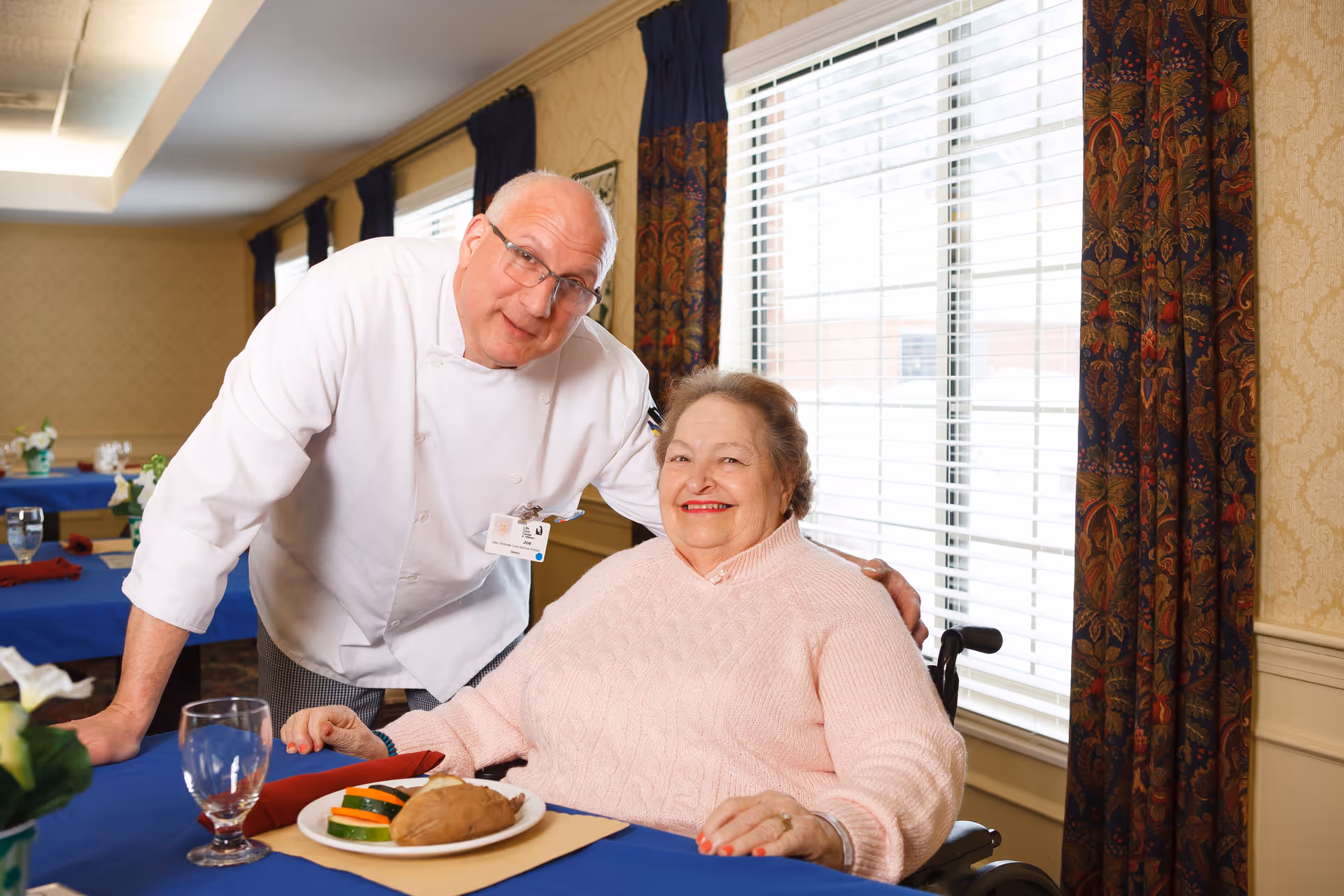 An elderly woman in a wheelchair wearing a pink sweater smiles while sitting at a dining table with a plate of food. A chef in a white uniform and glasses leans toward her with a friendly expression. The room has large windows with blinds and patterned curtains, and tables are set with blue tablecloths and glassware.