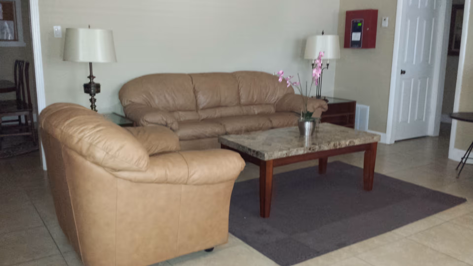 A living room area with two tan leather sofas arranged around a rectangular coffee table with a marble top. There is a potted orchid plant on the coffee table. Two table lamps with white shades are placed on side tables behind the sofas. The room has tiled flooring and beige walls, with a white door and a red fire alarm box visible in the background.