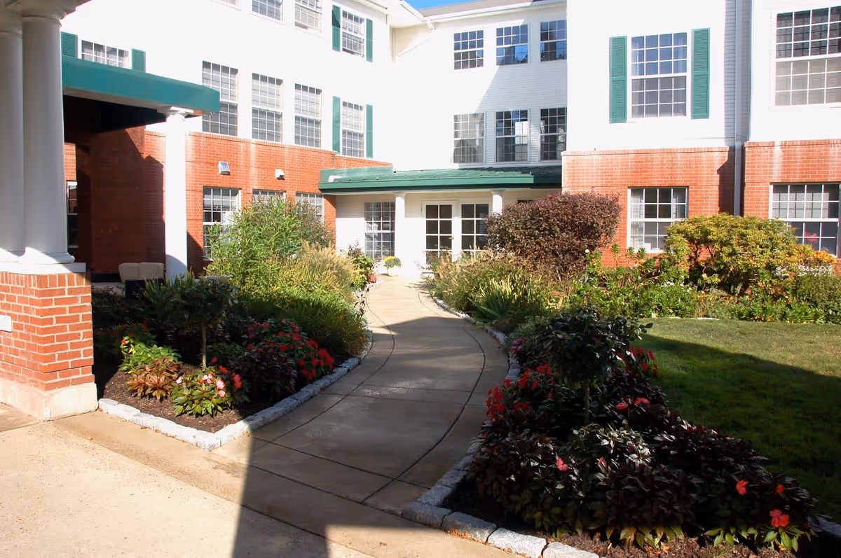 Outdoor walkway leading to the entrance of a senior living facility building with red brick and white siding exterior, green shutters, and landscaped garden beds with various plants and flowers on either side of the path.