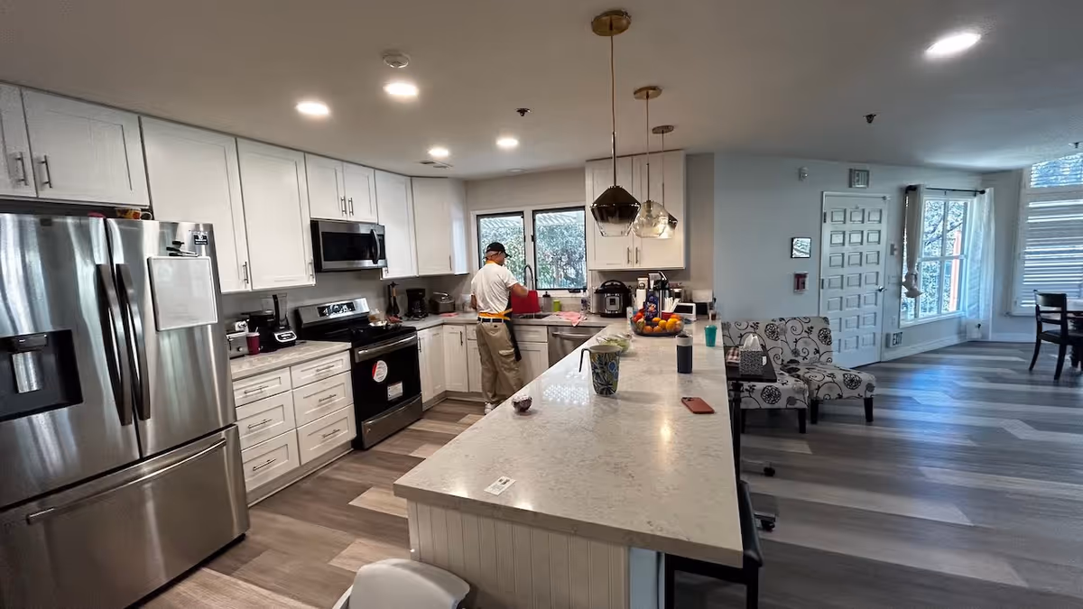 A modern kitchen with white cabinets, stainless steel refrigerator and oven, and a long marble countertop island. A person is standing near the sink washing dishes. The kitchen opens into a living area with patterned chairs and large windows letting in natural light.