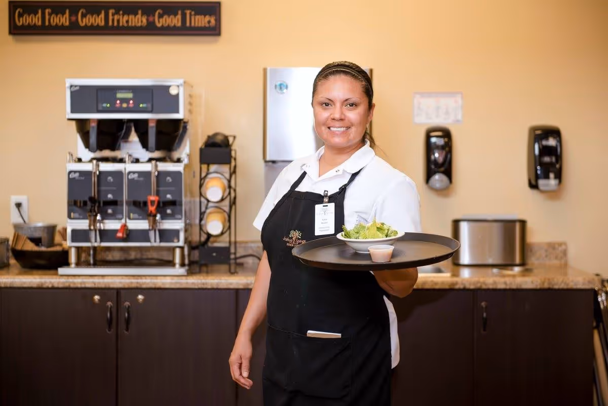 A smiling female staff member wearing a black apron and white shirt holds a tray with a bowl of salad and a small container of dressing in a dining or food service area. Behind her is a coffee machine, a rack with cups, and a countertop with various kitchen items. A sign above reads 'Good Food • Good Friends • Good Times'.