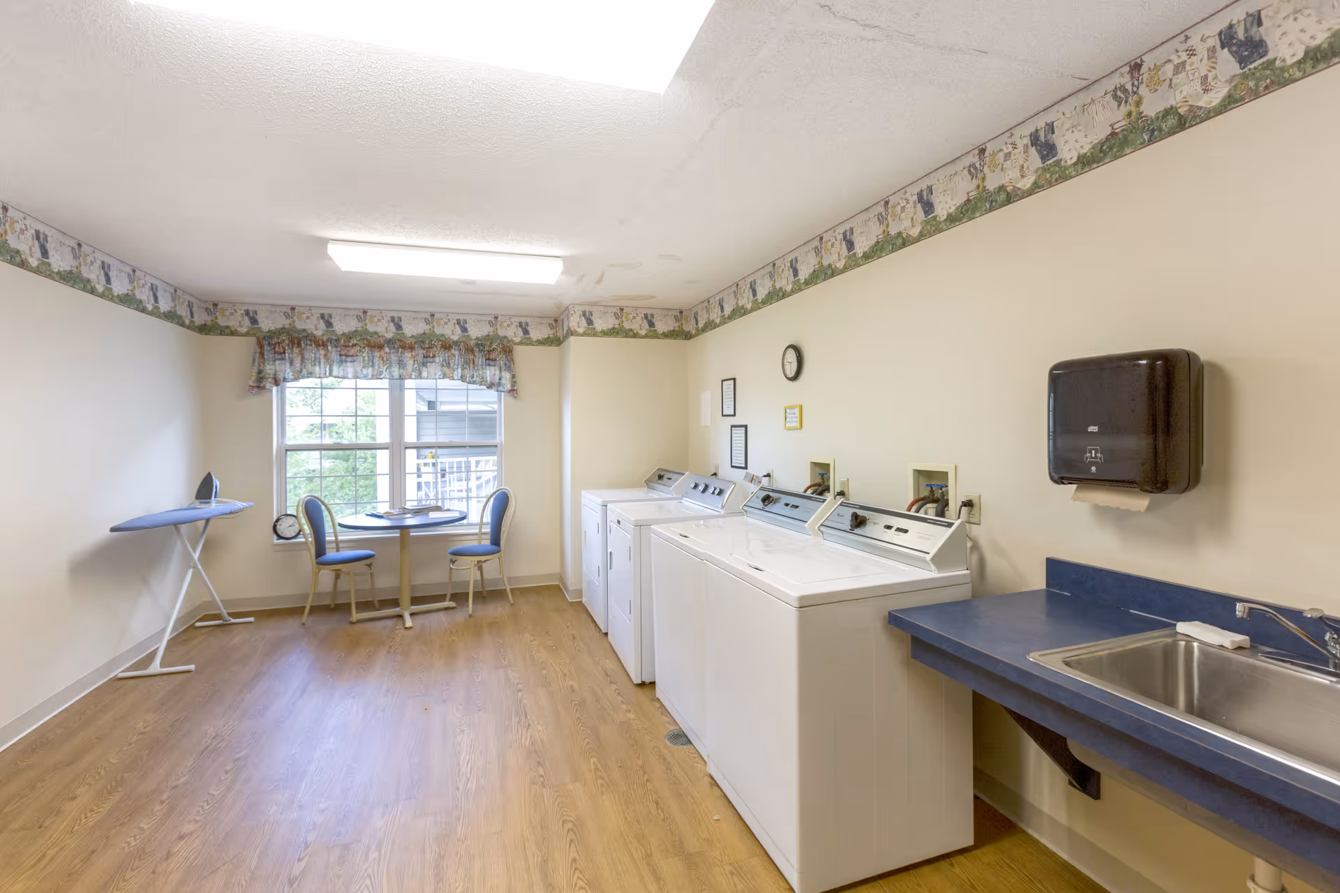Laundry room with three white washing machines and dryers along the right wall, a blue countertop with a sink, a paper towel dispenser above the sink, an ironing board on the left side, and a small round table with two chairs near a window with floral curtains.