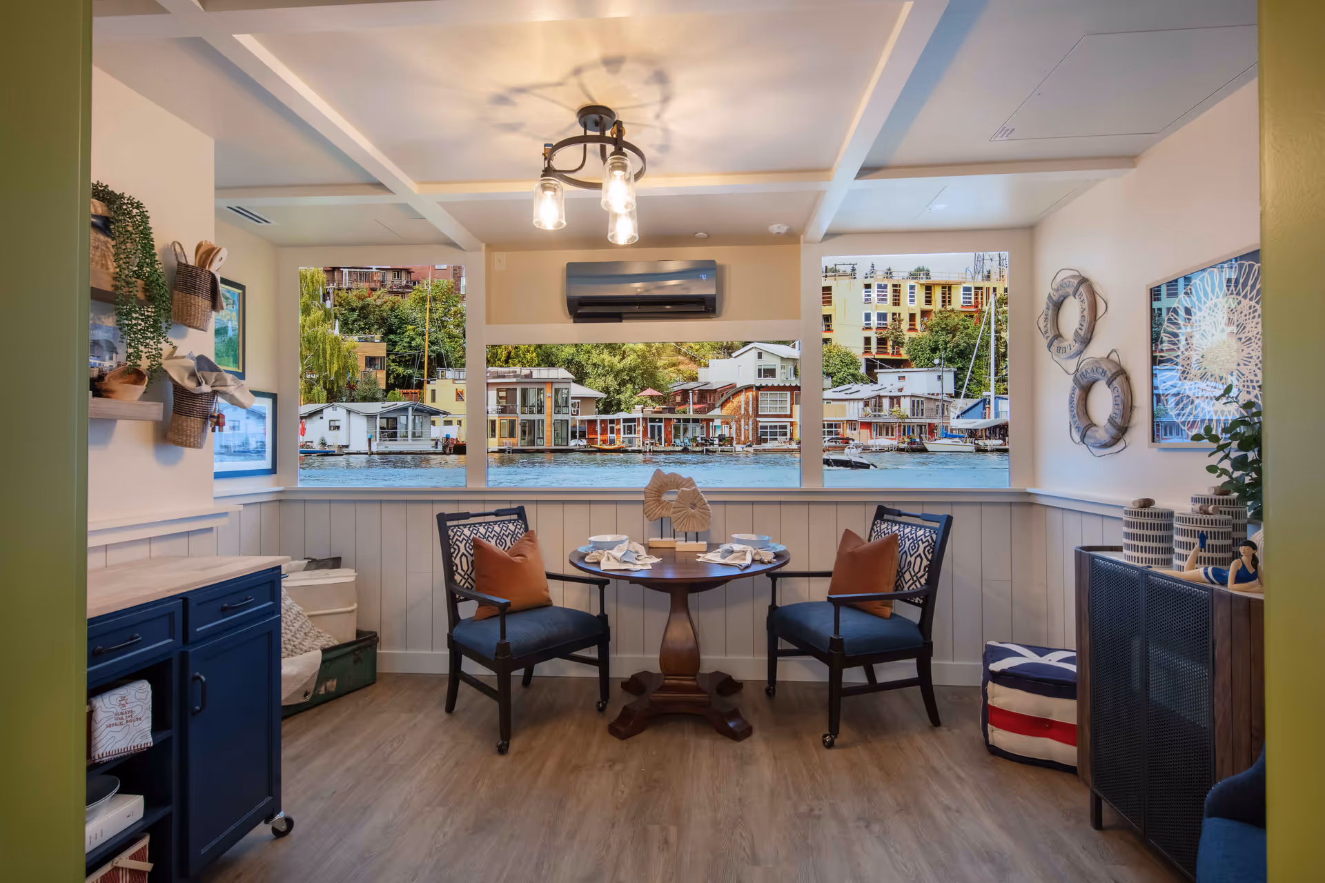 A cozy dining nook with a round wooden table set for four, two blue cushioned chairs with patterned backs and brown pillows, and large windows showcasing a waterfront view with houseboats and greenery. The room features light wood flooring, white paneled walls, nautical decor including life rings, and a ceiling light fixture with three bulbs.