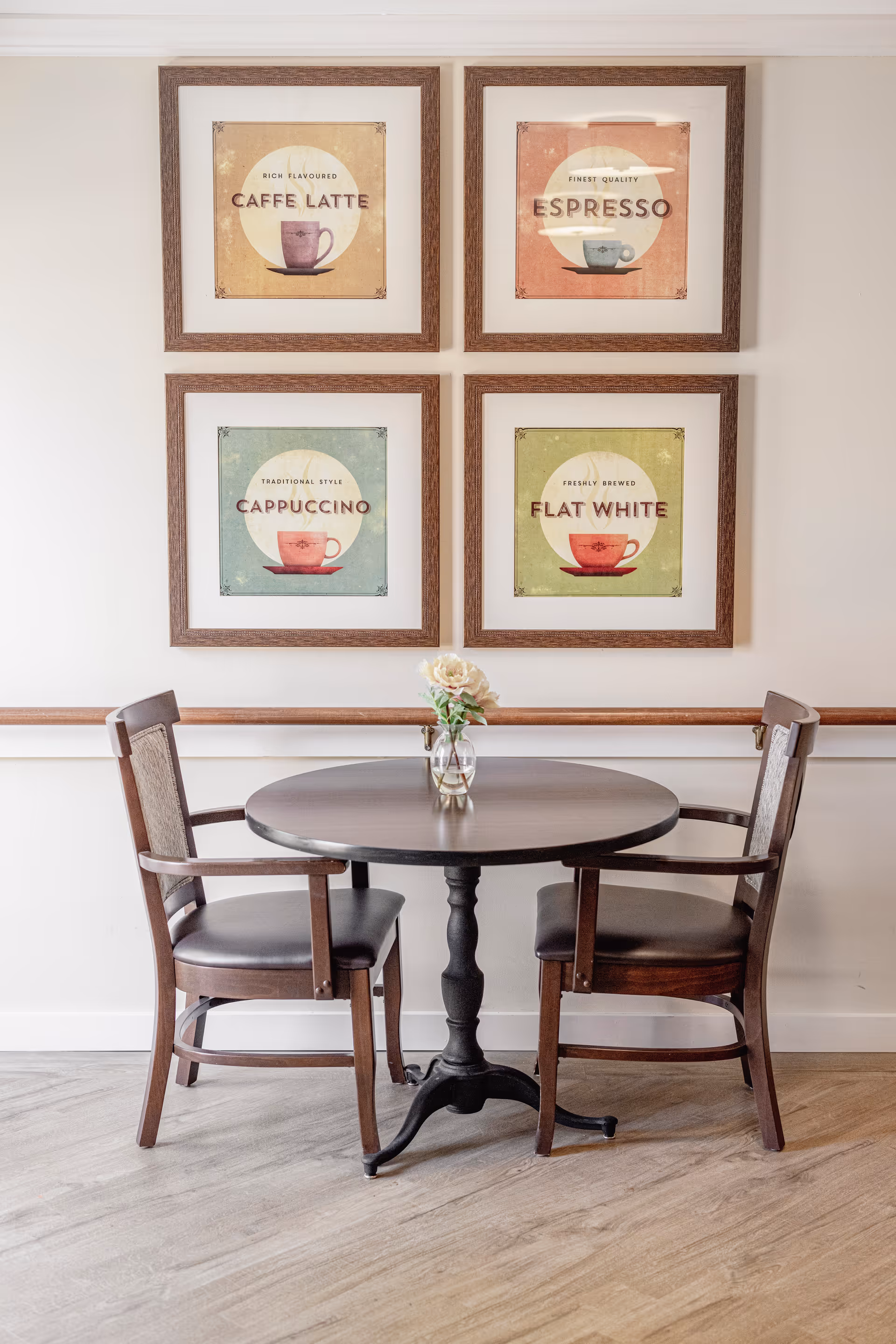 A small round wooden table with a vase holding a single flower, flanked by two wooden chairs with cushioned seats. On the wall behind the table are four framed pictures, each depicting a different coffee type: Caffe Latte, Espresso, Cappuccino, and Flat White.