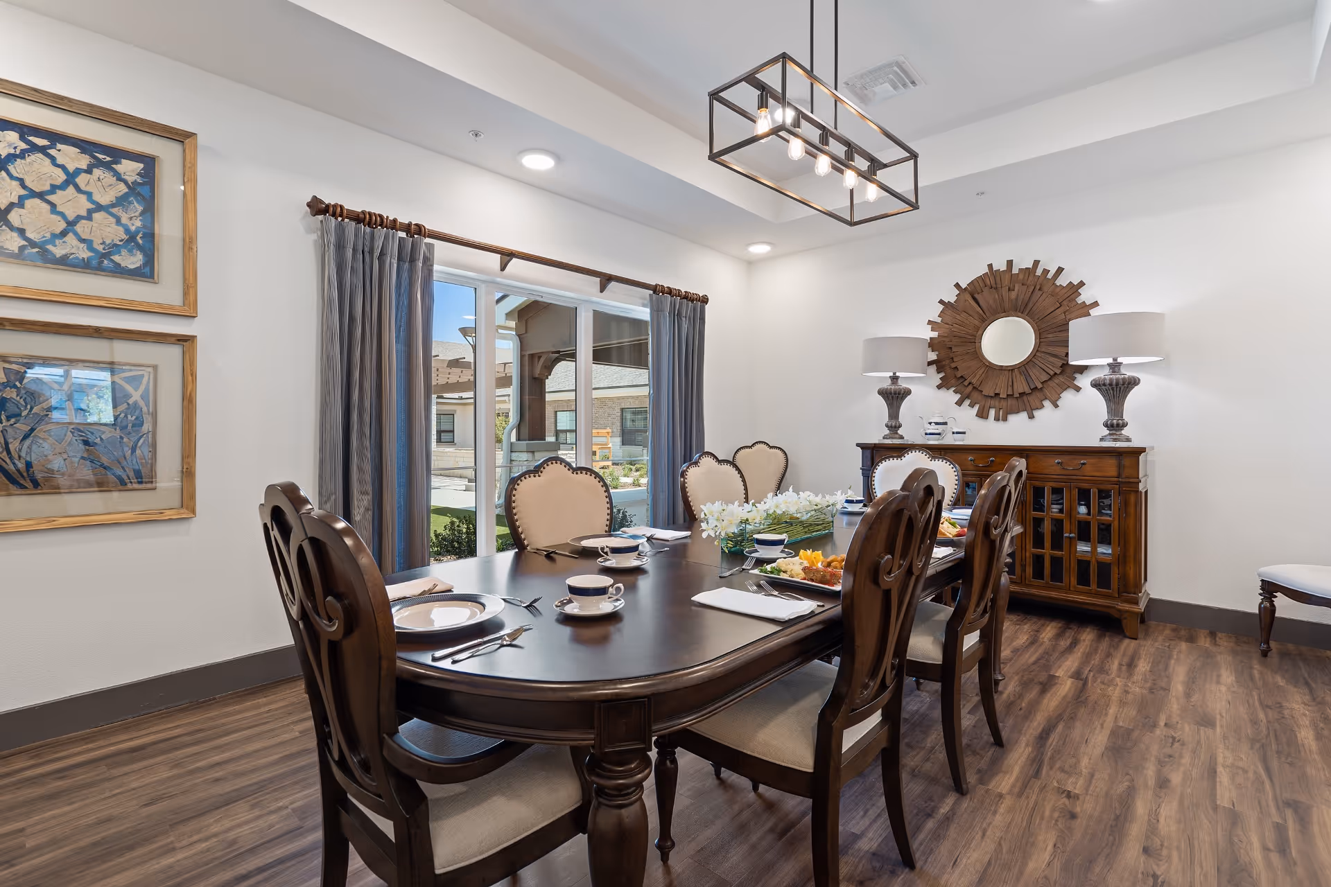 A well-lit dining room with a dark wooden dining table set for six, featuring beige cushioned chairs. The table has plates, cups, cutlery, and a floral centerpiece. There is a large window with curtains showing an outdoor view. On the wall, there are two framed artworks and a decorative wooden mirror above a wooden sideboard with two lamps.