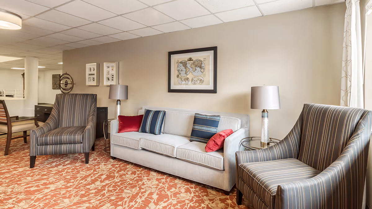 A cozy seating area in a retirement community featuring a light gray sofa with four decorative pillows in red and blue, flanked by two striped armchairs. There are two table lamps on glass side tables next to the sofa. The wall behind the sofa has a framed world map and two smaller framed pictures. The floor is covered with a patterned orange and beige carpet, and a window with light curtains is visible on the right side.