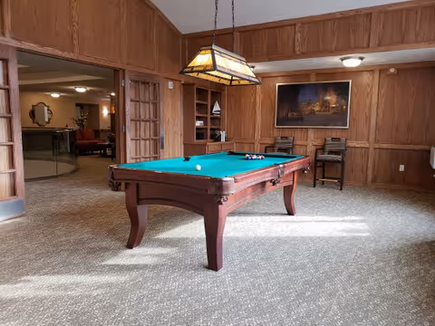 A room with wood-paneled walls featuring a pool table with billiard balls set up for a game. Above the table hangs a rectangular stained glass light fixture. In the background, there are two wooden chairs against the wall beneath a framed painting. An open doorway leads to another room with seating and a round mirror on the wall.