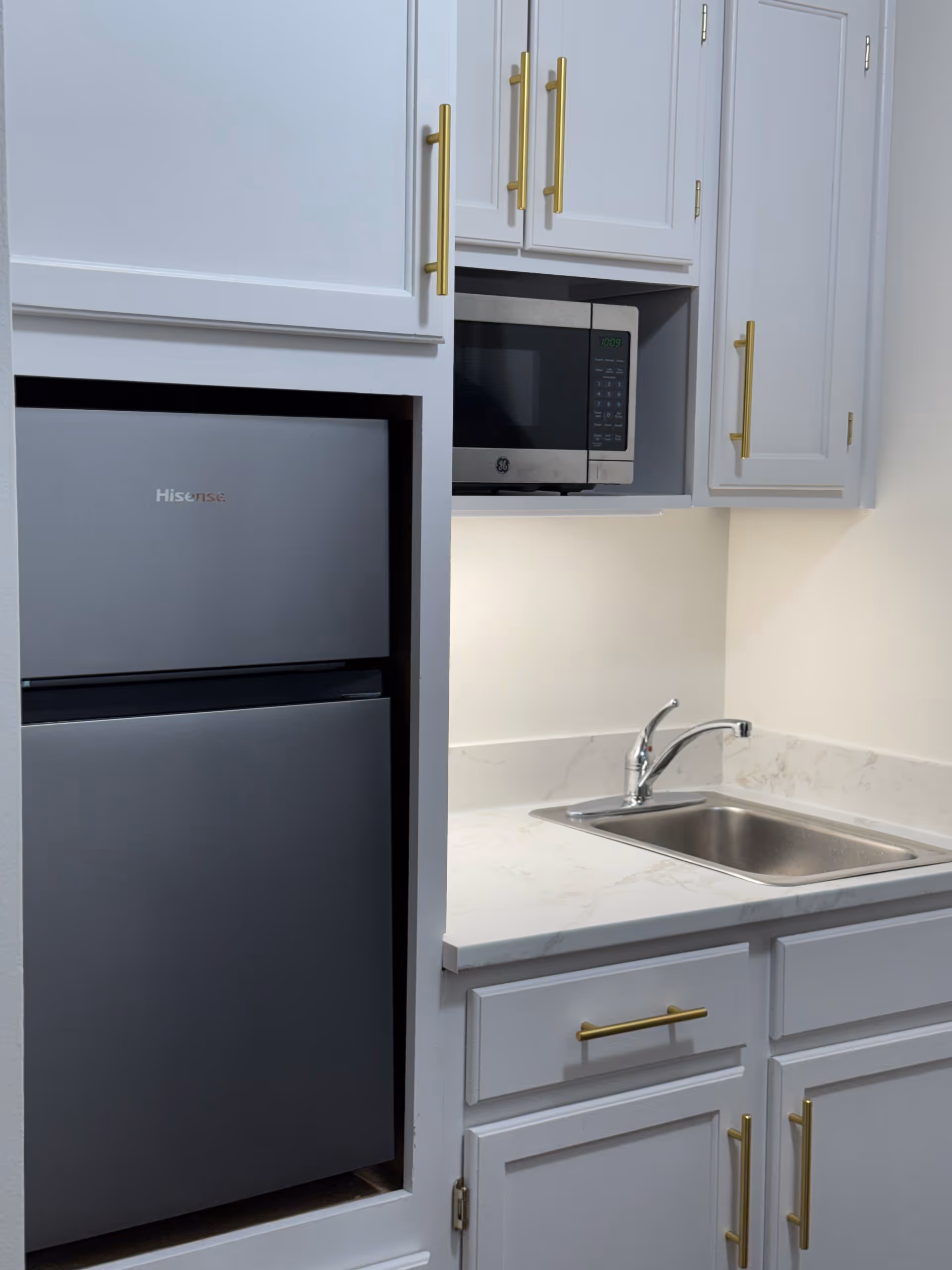 Small kitchenette with a Hisense mini refrigerator, built-in microwave, stainless sink, and white cabinets with gold handles.