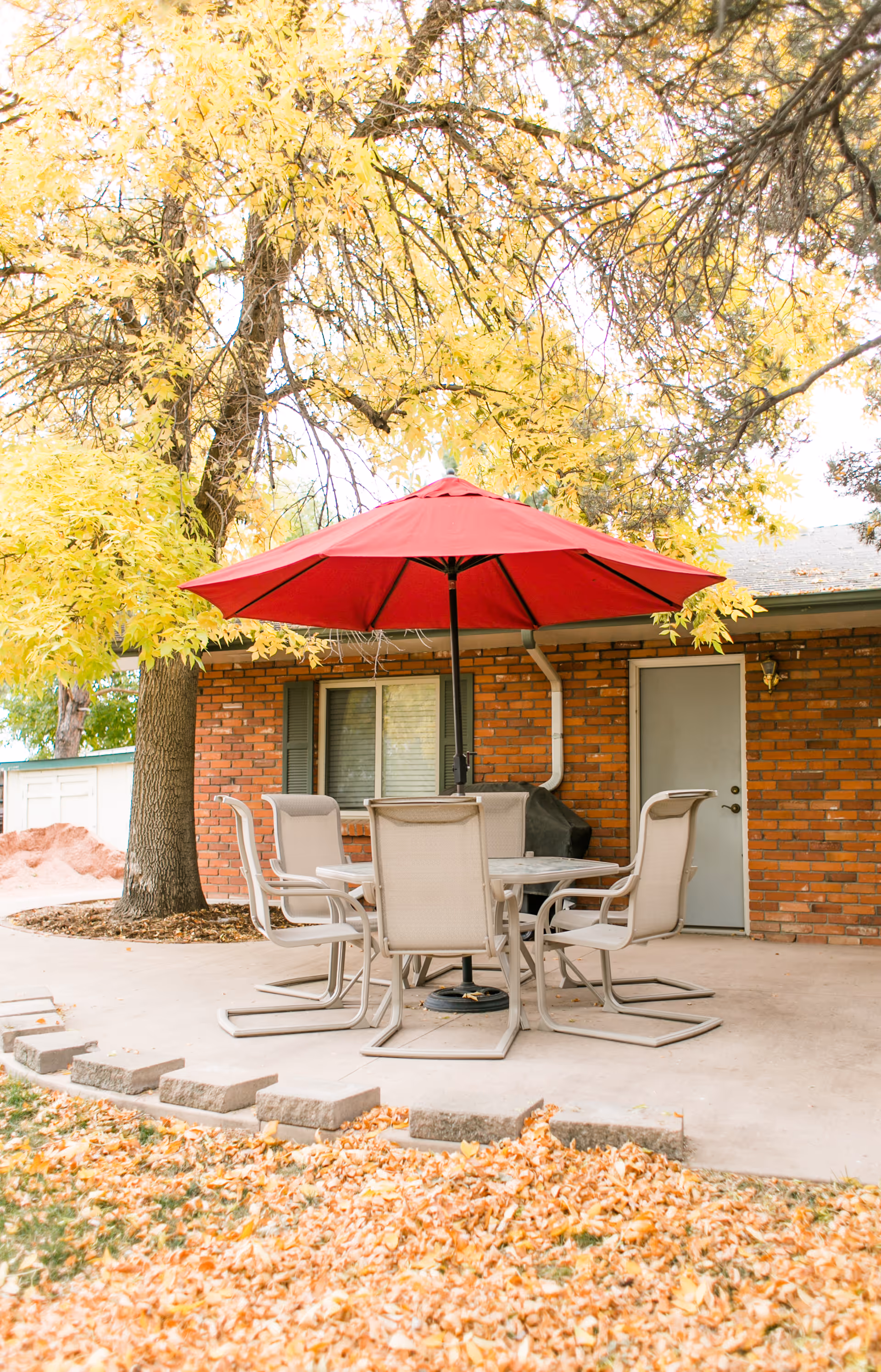 Outdoor patio with a round table, six chairs and a red umbrella in front of a brick building surrounded by autumn leaves.