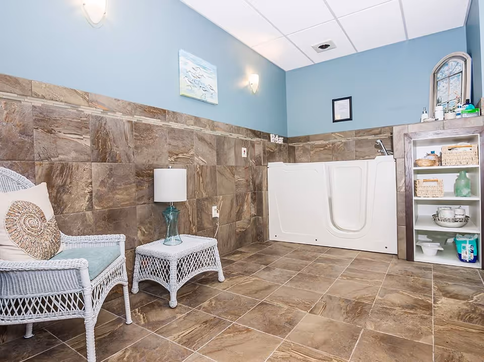 A bathroom with brown tiled walls and floor, featuring a white walk-in bathtub with a faucet and a small shelf unit holding various toiletries. There is a white wicker chair with a cushion and a matching wicker side table with a lamp. The walls are painted light blue and decorated with a painting and a framed certificate.