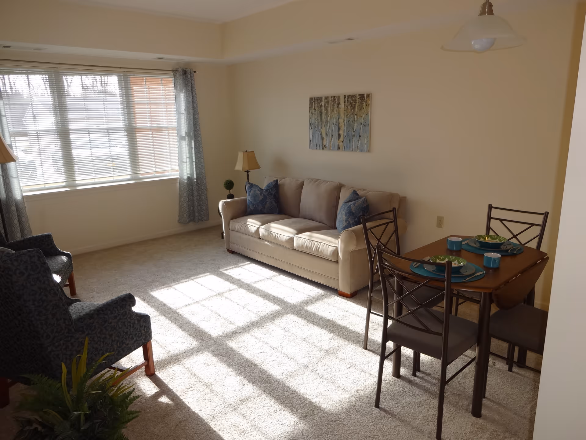 A bright living room with a large window letting in sunlight. The room features a beige sofa with two blue patterned pillows, two dark patterned armchairs, a small wooden dining table set for two with blue plates and cups, a floor lamp, a small plant, and a wall painting above the sofa.