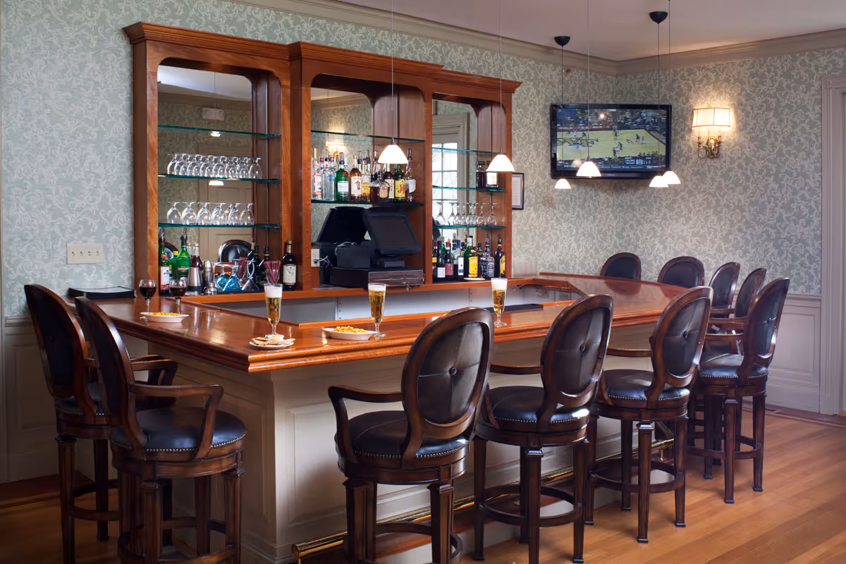Interior view of a bar area with a wooden counter and eight high-backed leather bar stools. Behind the bar, there are shelves with various bottles of liquor and glassware. Four glasses of beer and a glass of red wine are placed on the counter along with small plates of snacks. A flat-screen TV is mounted on the wall showing a sports game. The room has patterned wallpaper and wooden flooring.