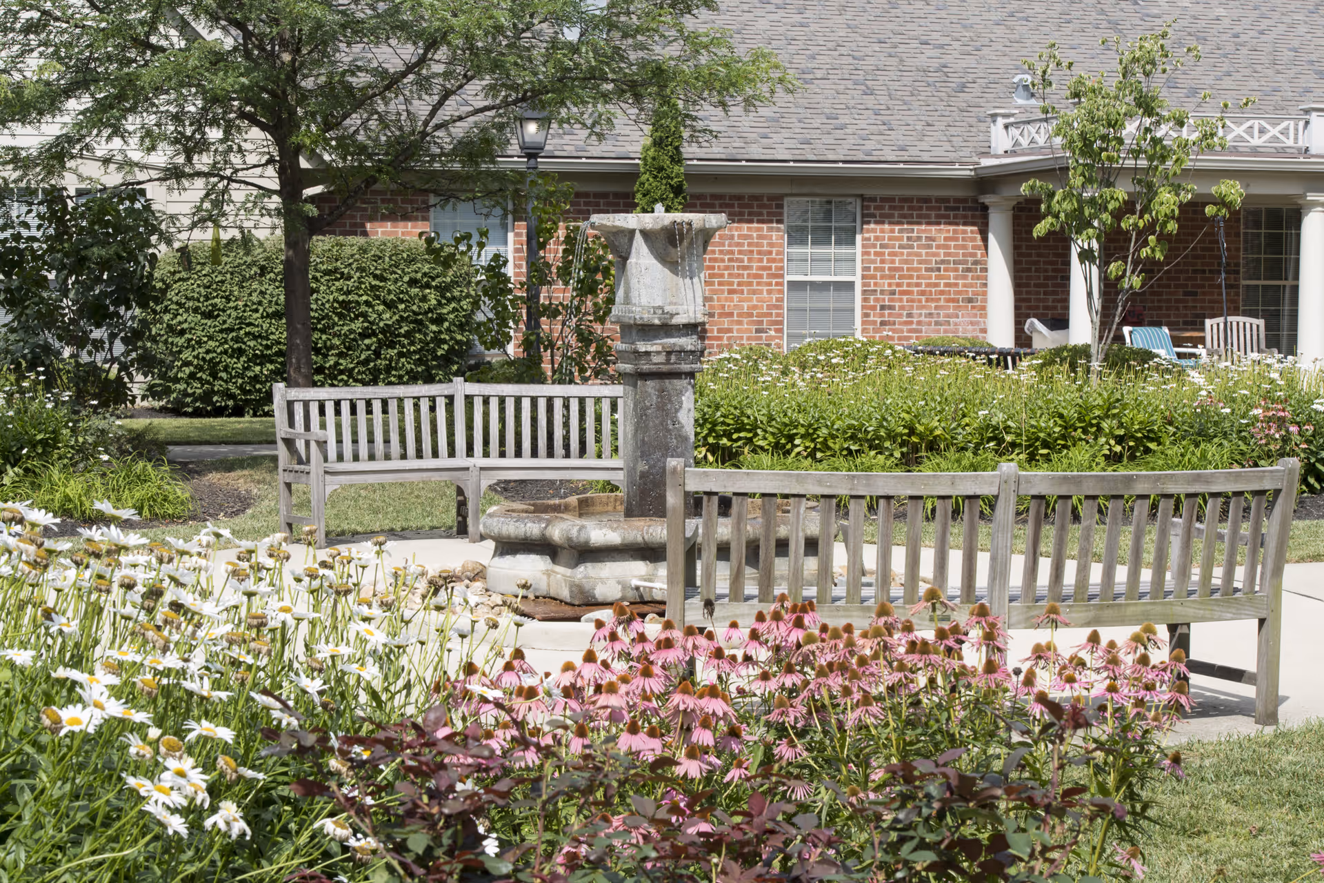 Outdoor garden area with wooden benches arranged around a stone water fountain. The garden features blooming white and pink flowers, green shrubs, and trees. In the background, there is a brick building with windows and a covered porch with chairs.