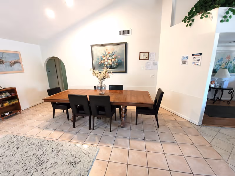 Dining area with a long wooden table and six chairs on a tiled floor beneath floral artwork.