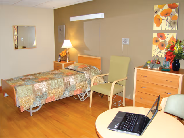 A neatly arranged bedroom in a senior living facility featuring a single bed with a colorful quilt, a wooden nightstand with a lamp and flowers, a green armchair, a wooden dresser with decorative items and flowers, two floral paintings on the wall, a round table with a laptop, and a mirror on the opposite wall.