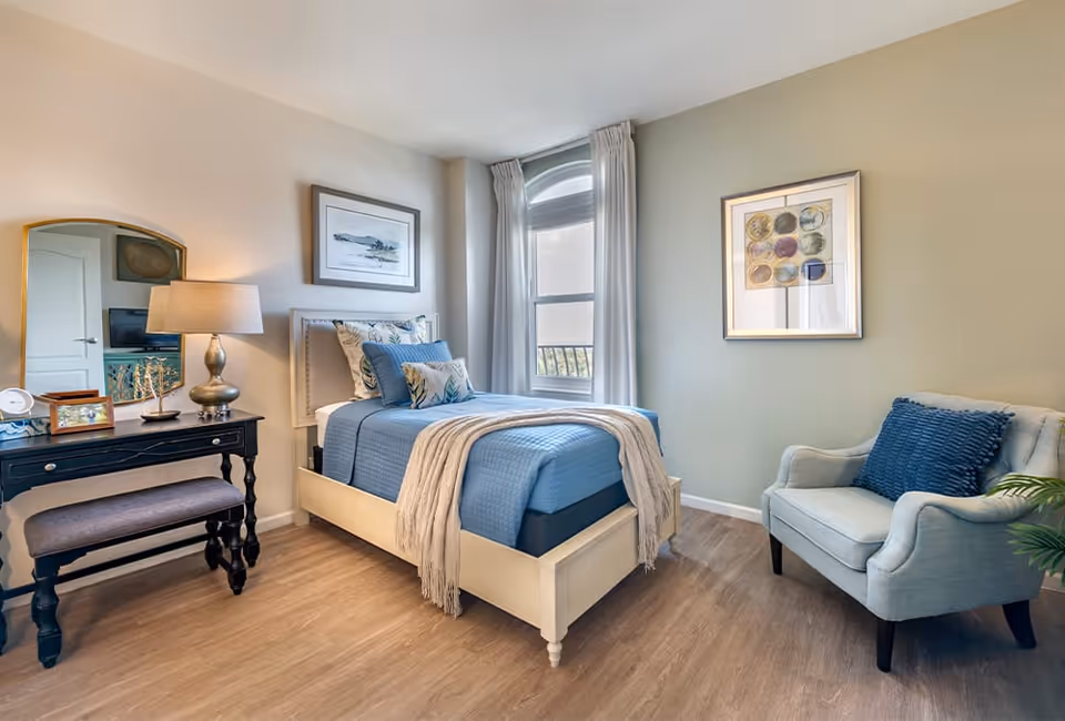 A bright, well-decorated bedroom with a single bed dressed in blue linens, a vanity table with a lamp, and an upholstered armchair.