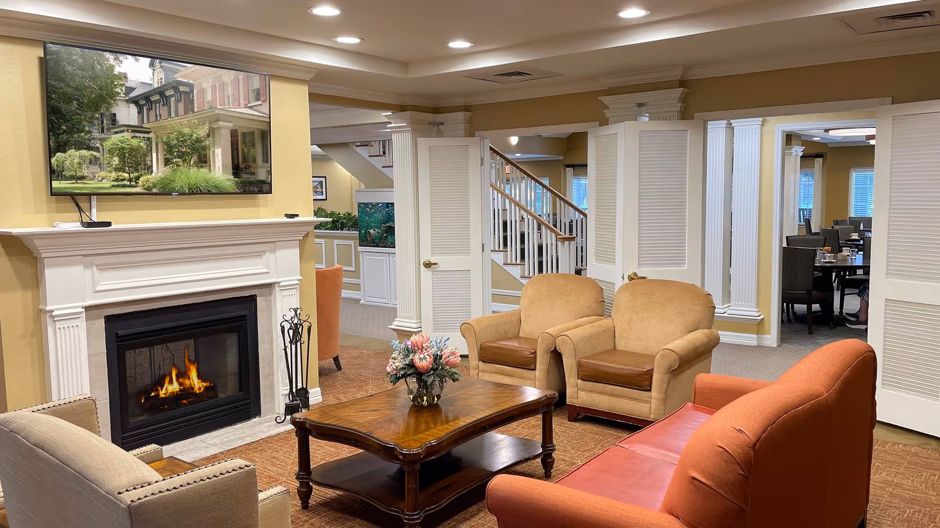 A cozy living room area in a senior living facility featuring a lit fireplace with a white mantel, a wooden coffee table with a floral arrangement, two beige armchairs, a red sofa, and a flat-screen TV mounted above the fireplace displaying a house exterior. The room has warm yellow walls, carpeted floors, and white columns and doors leading to a dining area in the background.