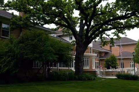 Brick senior living facility exterior with a large tree, lawn, and entrance.