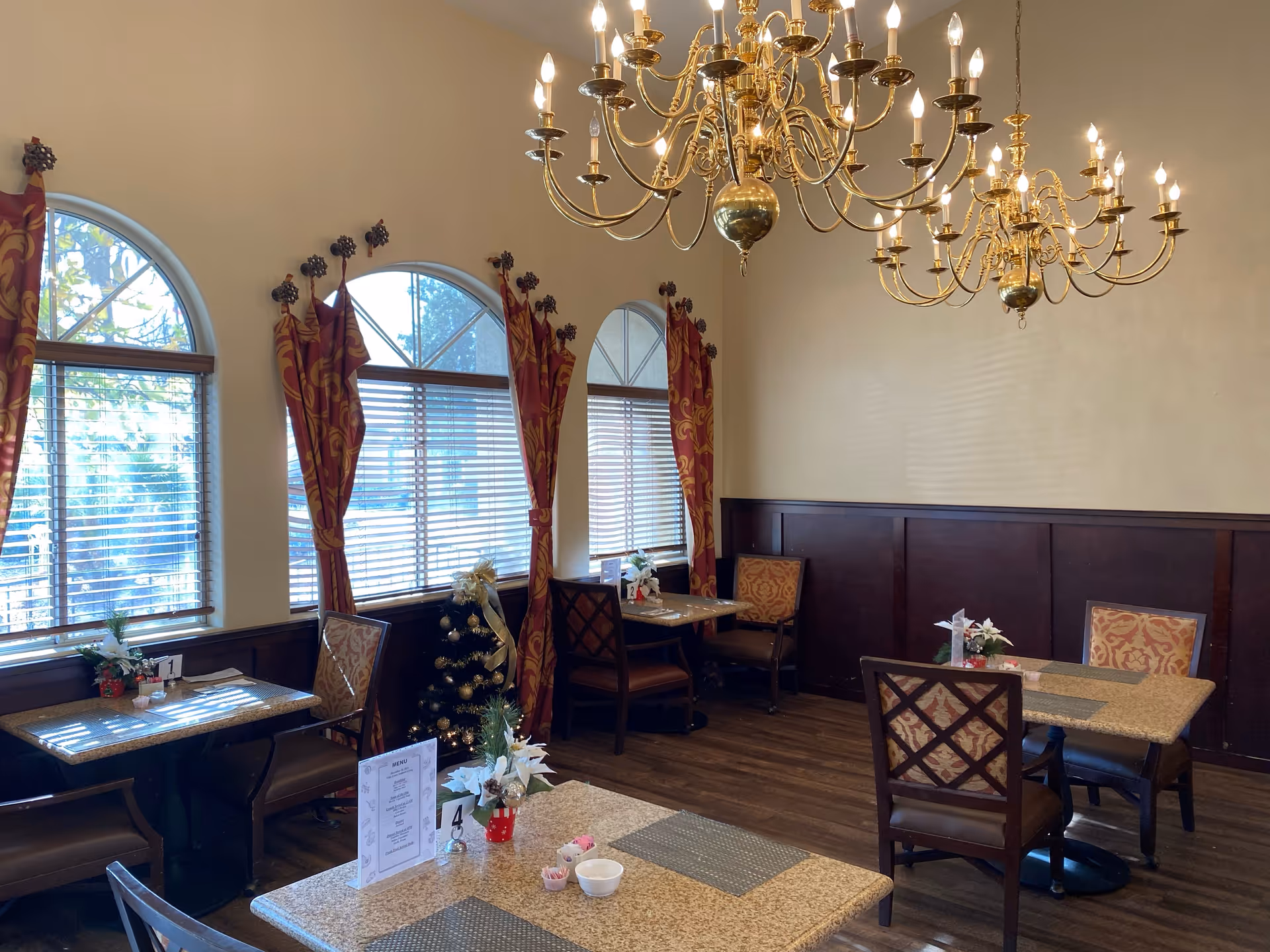 Dining room with several tables and chairs, decorated with small floral centerpieces. The room has large arched windows with red and gold patterned curtains, wooden paneling on the walls, and two ornate gold chandeliers hanging from the ceiling. A small decorated Christmas tree is visible near the windows.