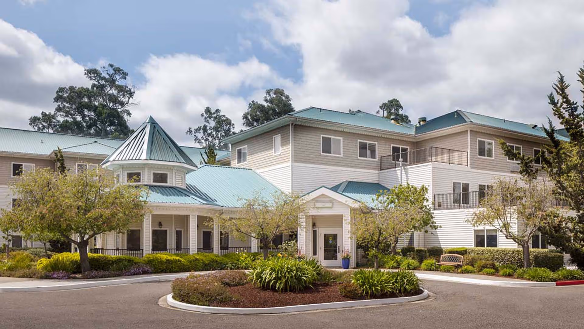 Exterior view of a senior living facility building with light-colored siding and teal metal roofs, surrounded by landscaped greenery and trees under a partly cloudy sky.