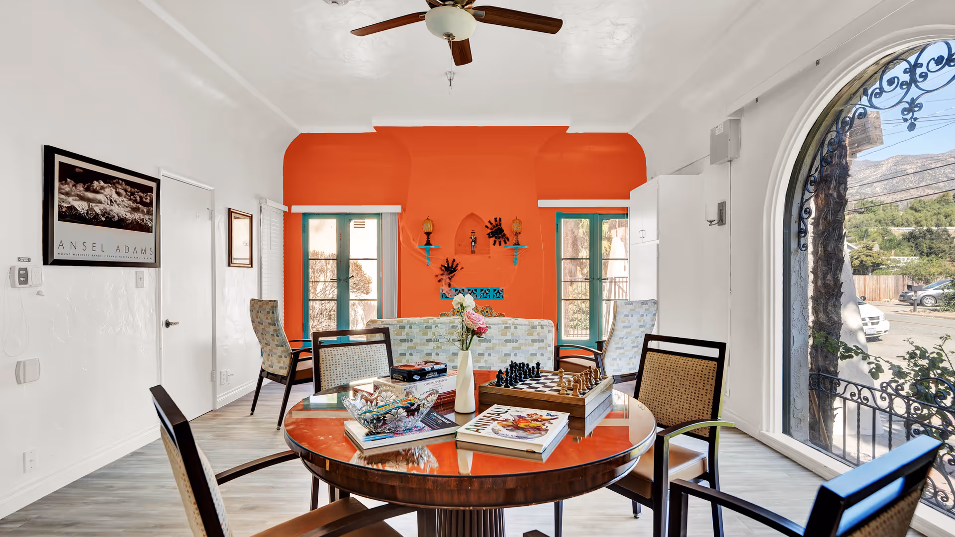 A bright and cozy living room area with a round wooden table in the foreground holding books, a chessboard, and a vase with flowers. Behind the table are cushioned chairs and a sofa with a colorful patterned fabric. The back wall is painted orange with two turquoise-framed windows and decorative shelves. A large arched window on the right side offers a view of the outdoors with trees and mountains visible. The ceiling has a wooden fan, and the walls are white with framed artwork.