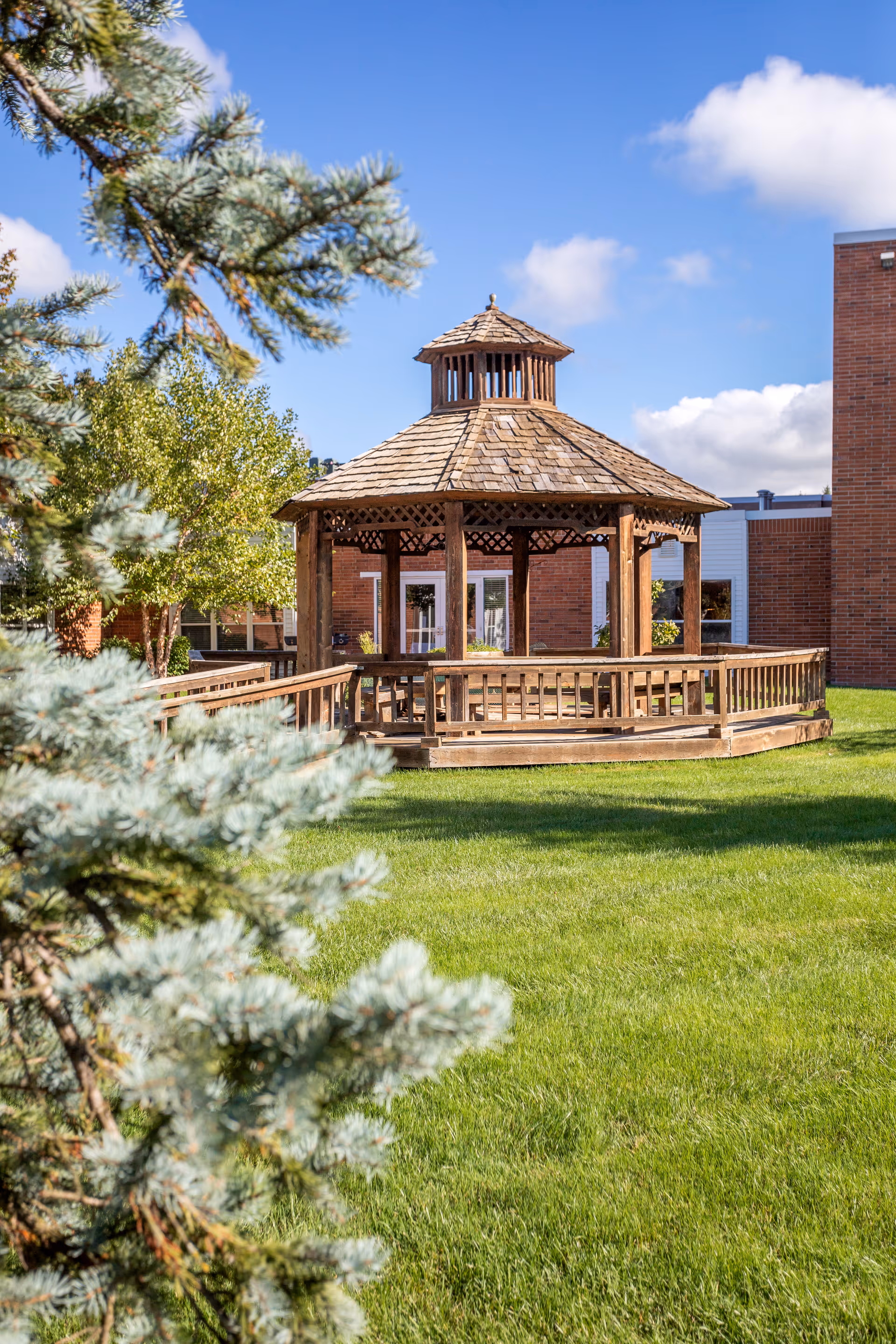A wooden gazebo with a shingled roof situated on a well-maintained green lawn, surrounded by trees and adjacent to a brick building under a blue sky with some clouds.