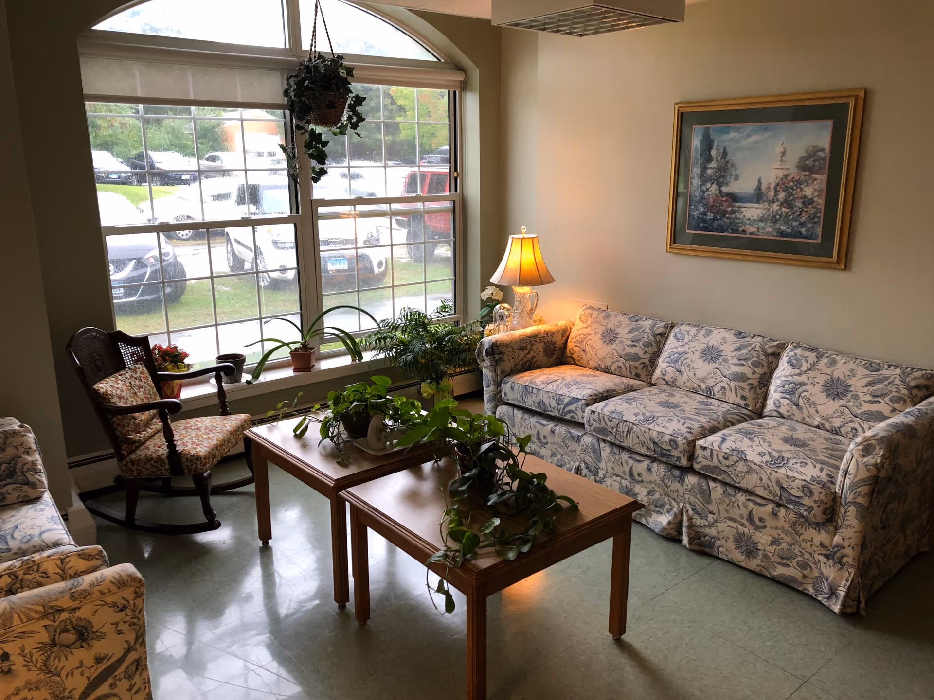 A cozy living room area with a floral-patterned sofa and matching armchair, a wooden rocking chair with a floral cushion, a wooden coffee table with green plants on it, a table lamp on a side table, and a large window with a hanging plant and a view of parked cars outside. A framed painting hangs on the wall above the sofa.