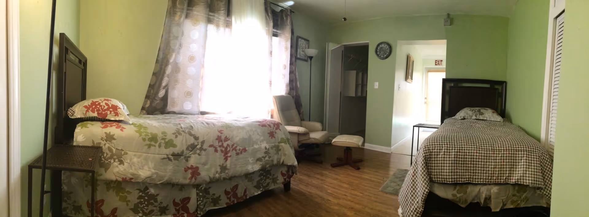 A shared bedroom with two single beds, a recliner and ottoman, floral and checkered bedding, light green walls, and wood flooring.
