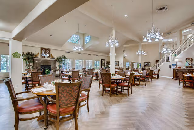 Spacious dining room with multiple wooden tables and chairs arranged neatly. The room features large windows allowing natural light, chandeliers hanging from the ceiling, a fireplace with decorative items above it, and a staircase leading to an upper floor.