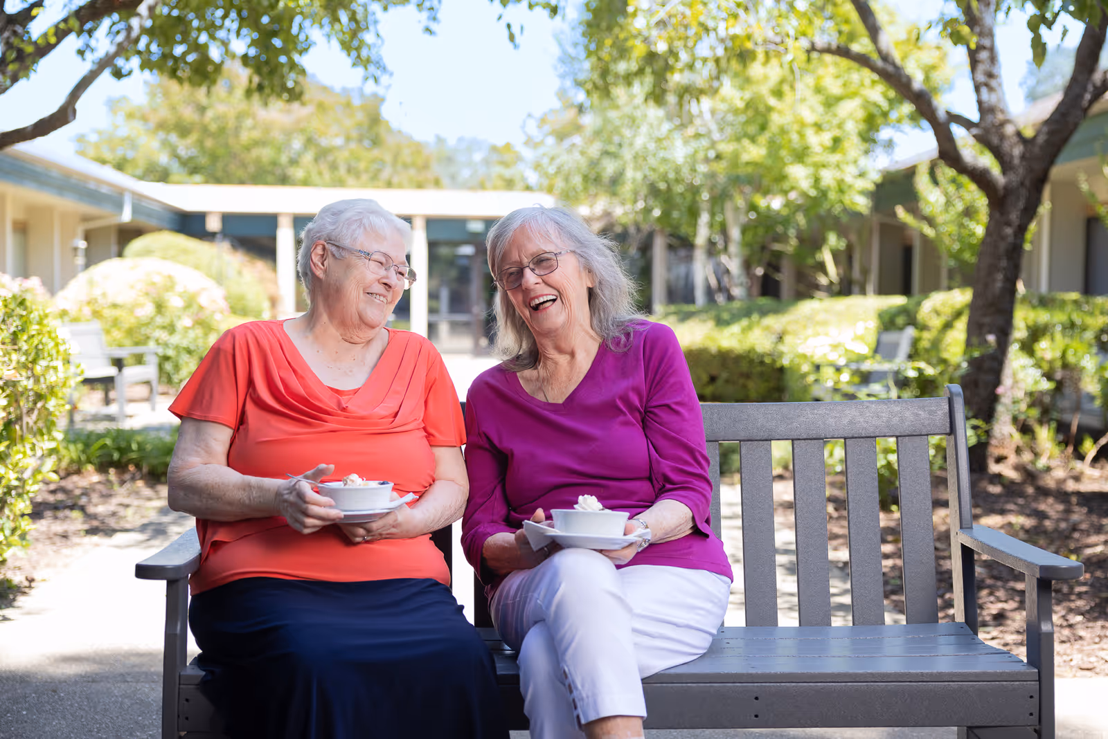 Two elderly women sitting on a wooden bench outdoors in a garden area of a senior living facility, smiling and holding bowls of dessert with whipped cream. Trees and bushes surround the paved pathway behind them.