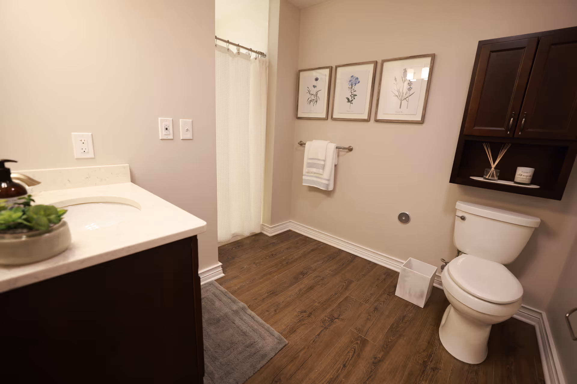 A clean and modern bathroom with a white toilet, a dark wood cabinet mounted above it, a white countertop with a sink and a small plant, a towel hanging on a rack, three framed botanical prints on the wall, a shower with a white curtain, and wood-look flooring.