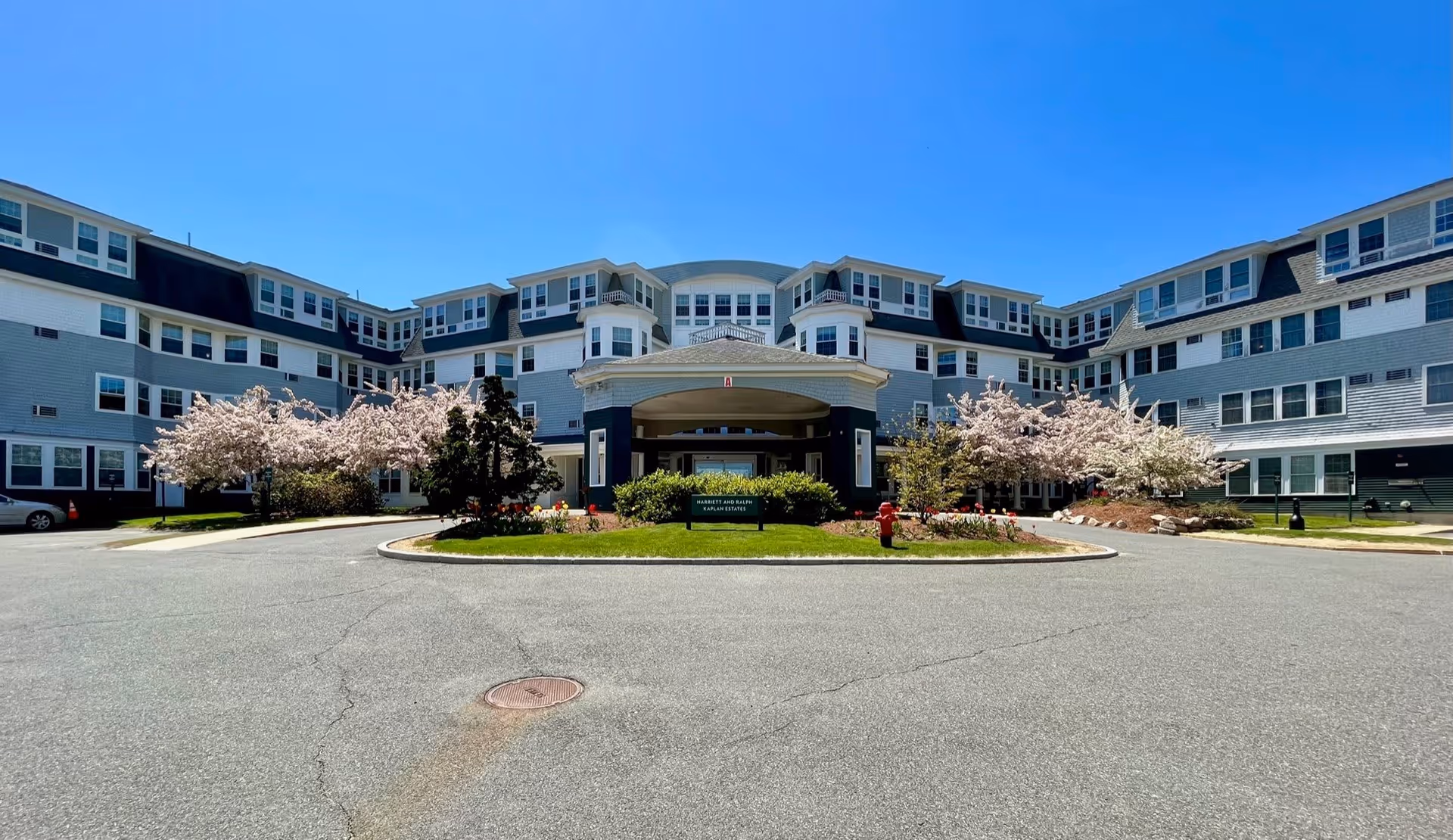 Front exterior view of Legacy at Harriett & Ralph Kaplan Estates, a large multi-story senior living facility with gray and white siding, multiple windows, and a covered entrance. The building is surrounded by blooming trees and landscaped greenery under a clear blue sky.