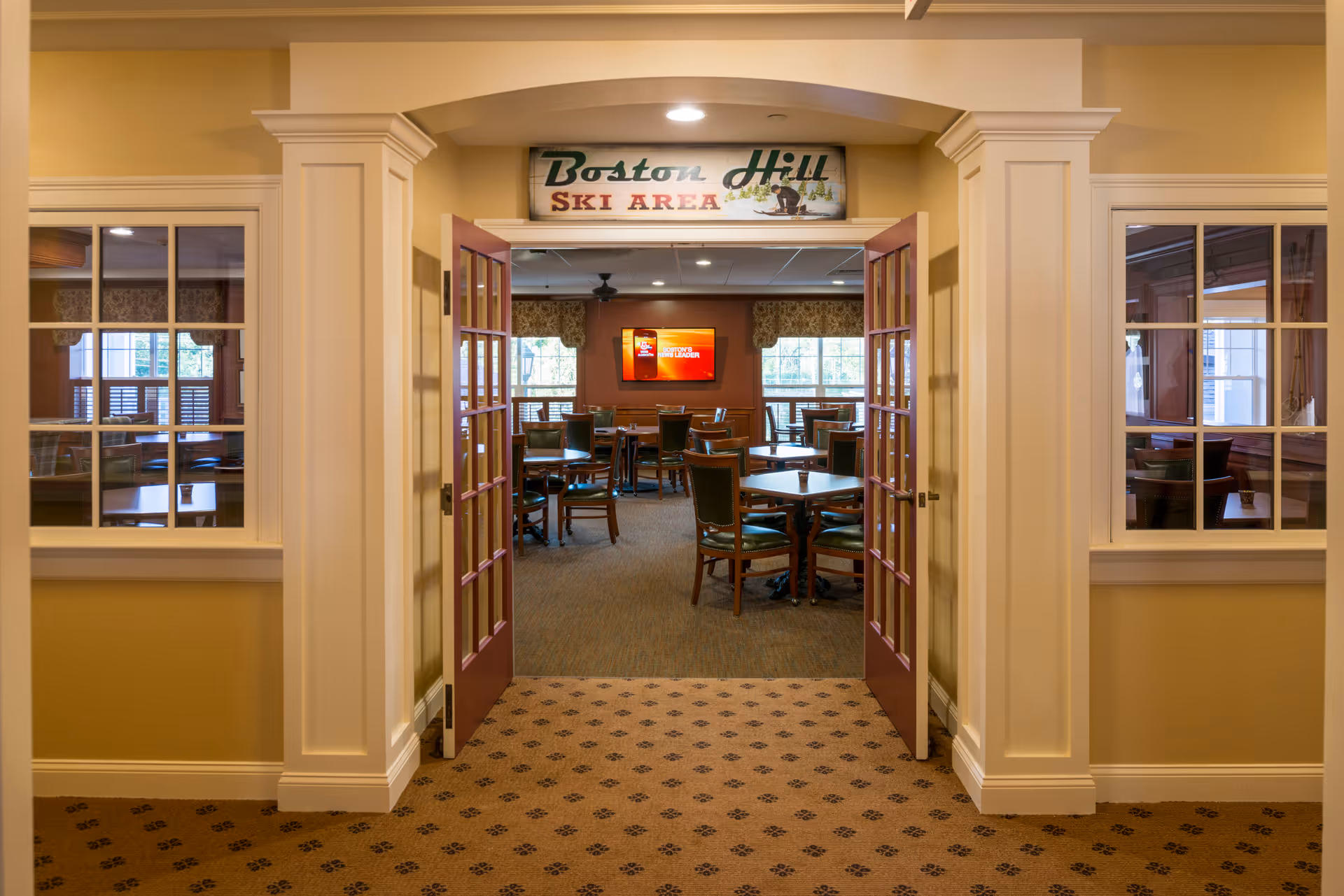 View through an open double door into a dining area with multiple tables and chairs. The walls are painted in warm tones, and there is a sign above the door that reads 'Boston Hill Ski Area'. Windows with curtains allow natural light into the room, and a television is mounted on the far wall.