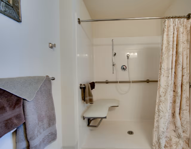 A bathroom shower area with a fold-down bench, grab bars, a handheld showerhead, and a floral-patterned shower curtain. Towels are hanging on a rack outside the shower.