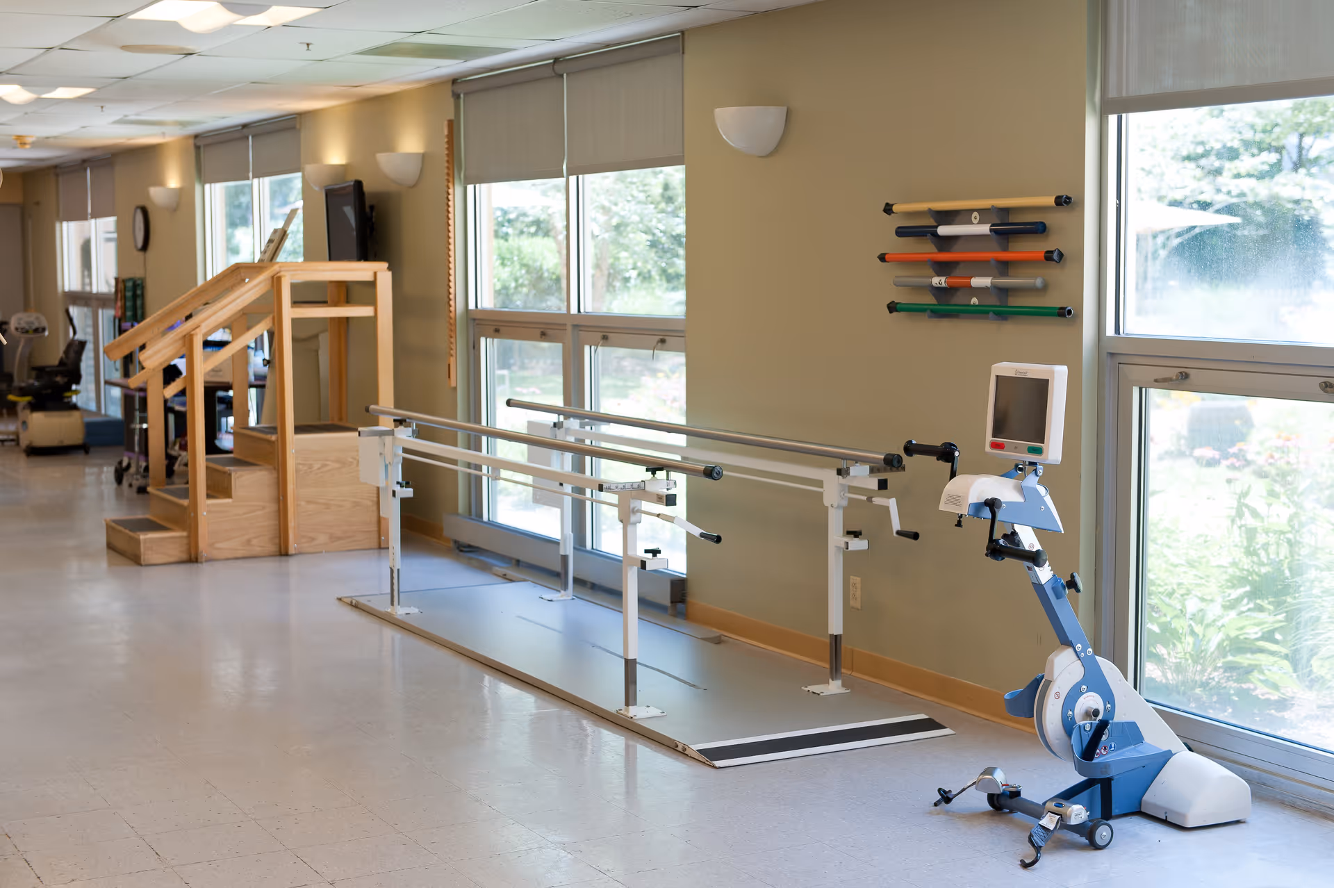 A bright rehabilitation room with parallel walking bars, a stationary rehab bike, and wooden practice stairs by large windows.