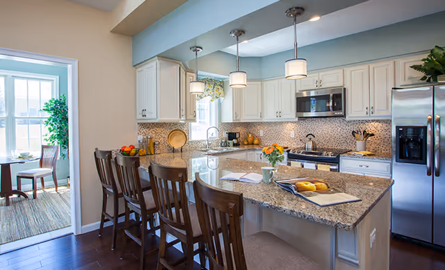 Bright kitchen with white cabinets, granite countertops, and stainless steel appliances including a refrigerator and microwave. Four wooden bar stools are lined up along the kitchen island, which has an open book, a cup, and a bowl of fruit on it. A doorway leads to a sunlit dining area with a table and chairs, and a large green plant is visible near the window.