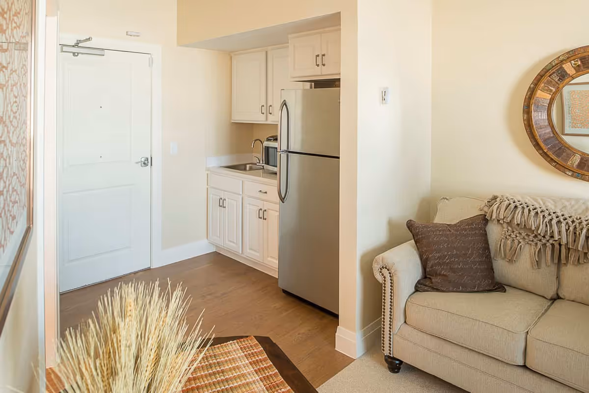 A cozy interior space featuring a small kitchen area with white cabinets, a stainless steel refrigerator, and a sink. Adjacent to the kitchen is a beige upholstered sofa with a decorative pillow and a textured throw blanket. A round wooden-framed mirror hangs on the wall above the sofa. The floor is a combination of wood and carpet, and a white door is visible in the background.