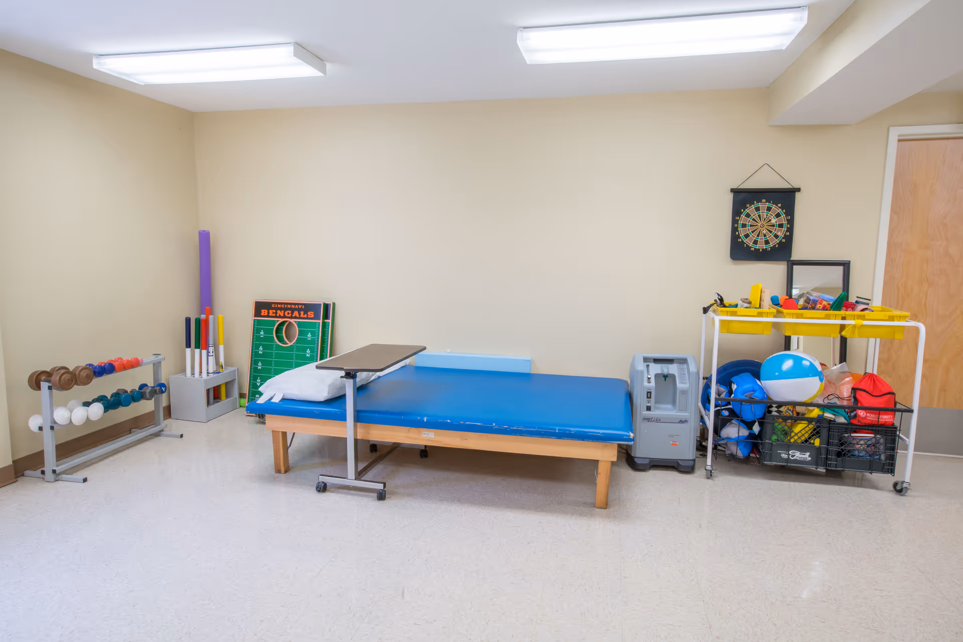 A beige therapy room with a blue padded treatment table, weights, exercise equipment, and a cart of therapy toys and supplies.